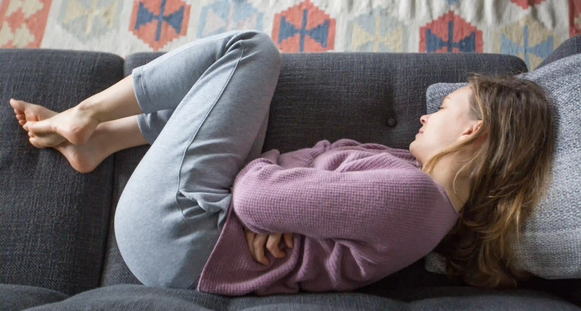 Mujer en posición fetal sobre un sofá con las manos en el abdomen, representando los síntomas físicos de la ansiedad y el malestar digestivo emocional.