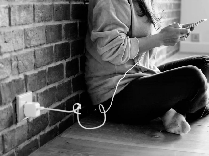 Persona sentada en el suelo usando un móvil mientras carga la batería, representando la dependencia digital y la necesidad de terapia para el estrés.